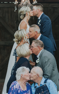 An elderly couple sharing a kiss while sitting on a porch railing, with a woman in a blue dress in front of them, and a man in glasses and a gray suit, all in a heartfelt moment.