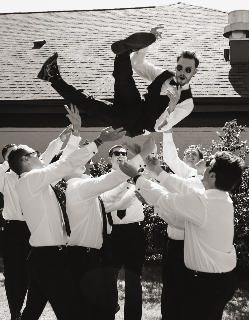 A group of five men in white shirts and sunglasses lifting a woman in a black outfit into the air outdoors. The woman is smiling, wearing sunglasses and a leather jacket, with her legs bent backward in mid-air.