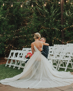 Bride and young child dancing outdoors at a wedding ceremony with white chairs and string lights in the background.