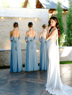 Rose Briar Place Four women in long blue dresses standing outdoors with one woman in a white dress, some trees, and a modern outdoor setting in the background.