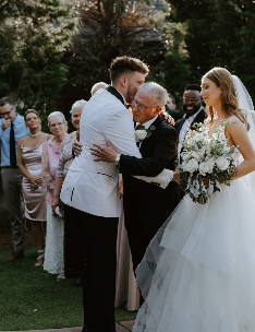A wedding ceremony outdoors with a bride and groom exchanging vows, surrounded by family and friends. The groom wears a white suit, the bride holds a bouquet, and the scene is set in a lush garden.