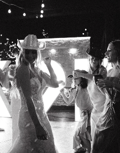 Group of women and children dancing together in a dimly lit setting, with one woman wearing a cowboy hat and the others holding drinks and wearing casual attire.