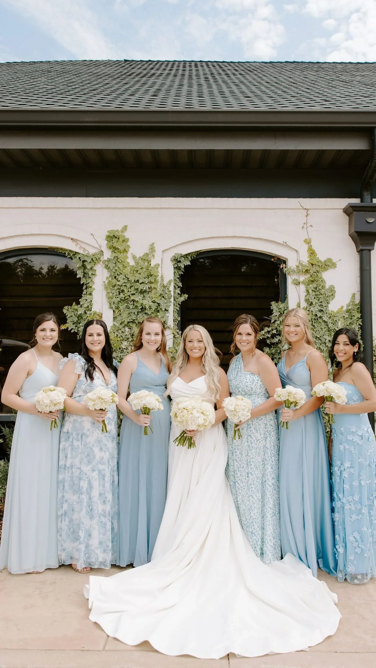 Group of seven women in pastel blue dresses, standing outdoors holding white bouquets, in front of a white building with green vines, during daytime.