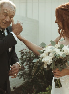 An elderly man and a woman in a wedding dress sharing a moment outdoors. The woman is holding a bouquet of white flowers and touching the man's face, who is smiling and touching his eye.