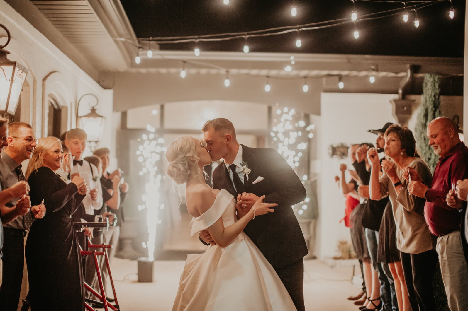 A bride and groom share a kiss during their wedding reception, surrounded by guests celebrating under string lights with sparklers and fireworks in the background.