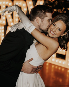 A couple dancing at a formal event, with the man in a tuxedo and the woman in a white dress, smiling and embracing each other. Bright lights and a decorated background are visible.