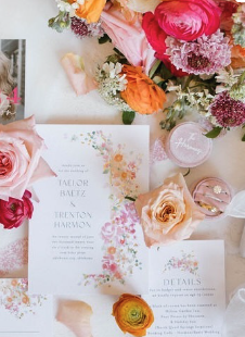 A wedding invitation surrounded by pink, orange, and purple flowers, a peach rose, and small candles.
