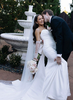 A bride and groom share a kiss outdoors beside a white fountain. The bride wears a strapless white wedding gown, holding a bouquet, with a veil. The groom wears a dark suit.