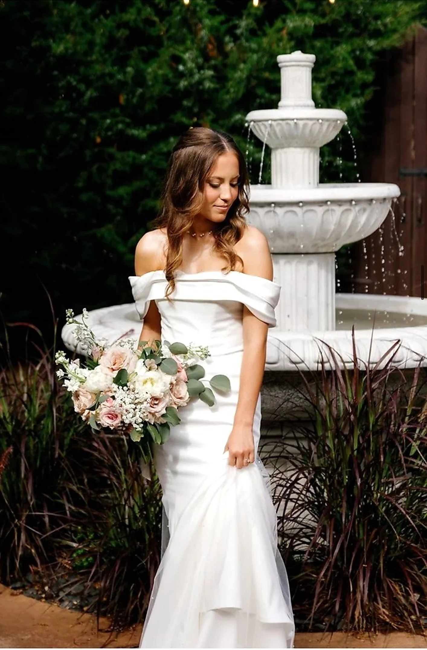A woman in a white off-the-shoulder wedding dress holding a bouquet of flowers stands outdoors near a white fountain, with dark greenery in the background.