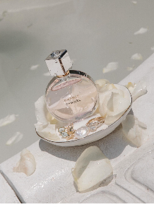 A clear perfume bottle lying in an open seashell, surrounded by white rocks on a white surface.