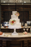 A white three-tiered wedding cake with floral decorations on a cake stand, set on a wooden table with champagne glasses in the background.