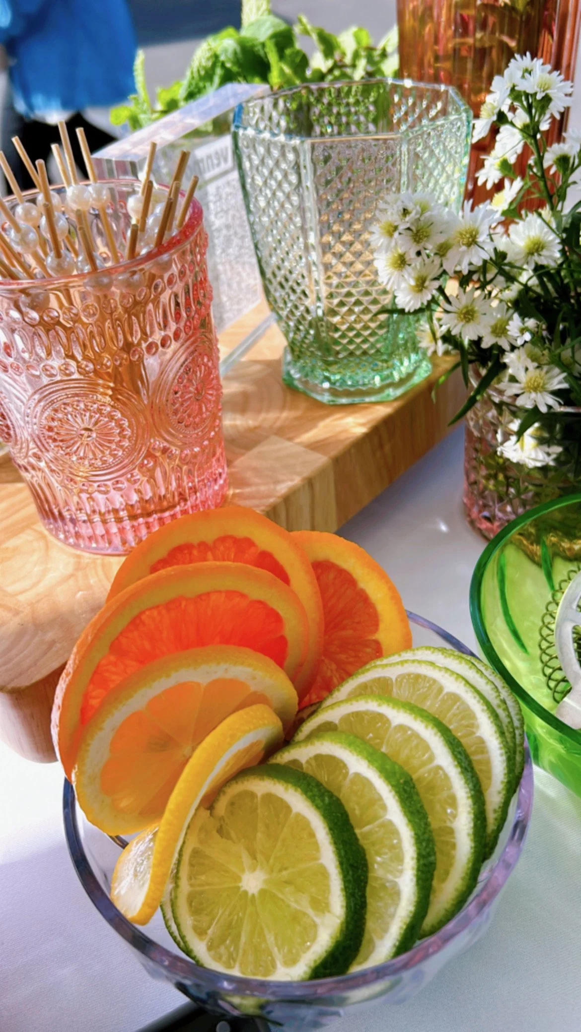 Slices of lemon and lime arranged in a glass bowl with pink and orange slices in the background, near decorative glassware and a vase with white flowers on a table.