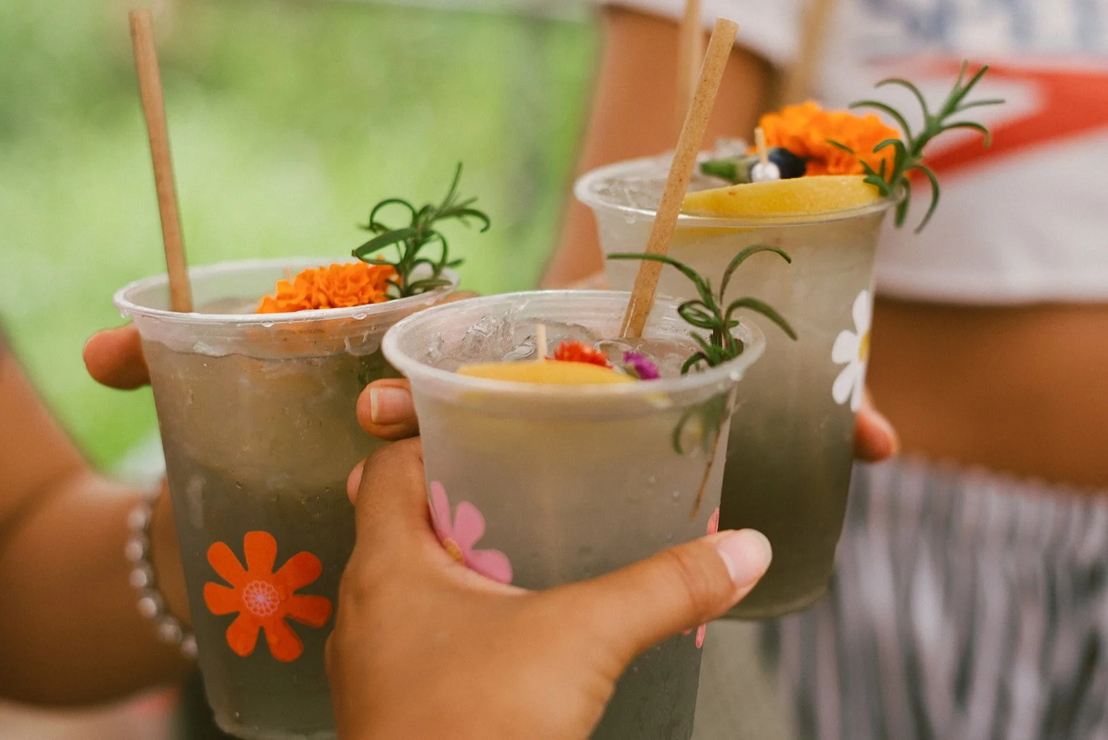 Three people holding drinks in clear cups decorated with flowers, garnished with marigold flowers, green herbs, and straws, toasting in celebration.