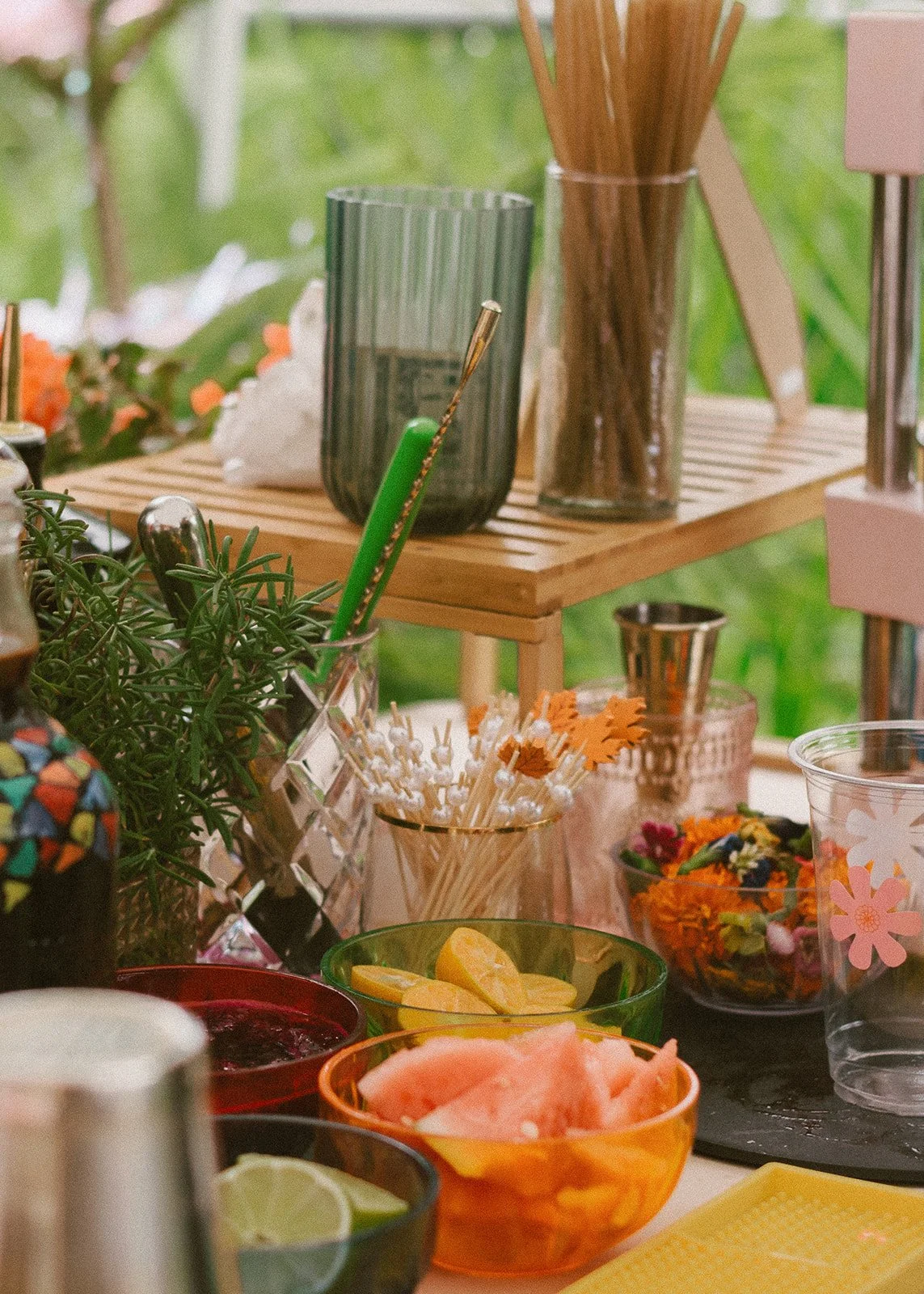 Colorful bowls of sliced lemons, watermelon, and lime on a table with various drink and fruit garnishes, with a background of plants and flowers.