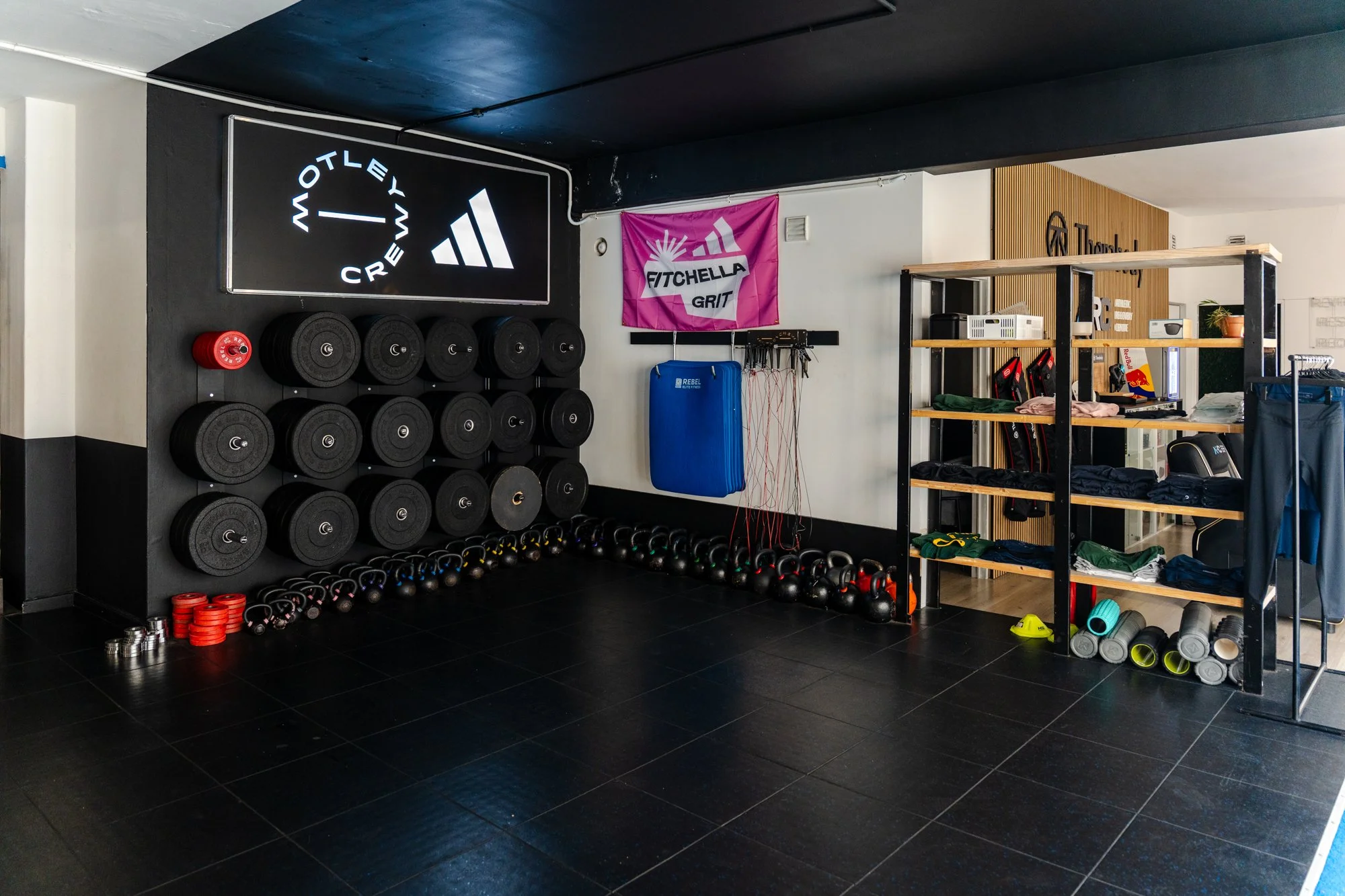 Fitness gym with black wall-mounted weight rack holding various black weight plates and red weight discs, kettlebells beneath, pink 'Fitchella Grit' banner, and metal shelves with workout gear, mats, and small appliances.