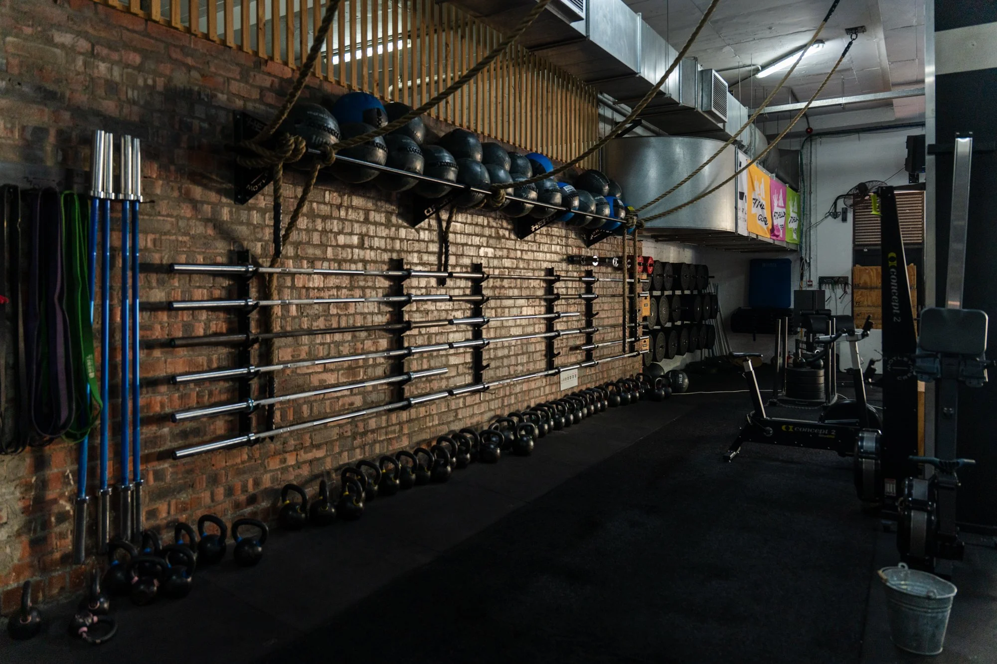 Interior of a gym with various workout equipment including barbells, dumbbells, kettlebells, medicine balls, resistance bands, and ropes against an exposed brick wall.