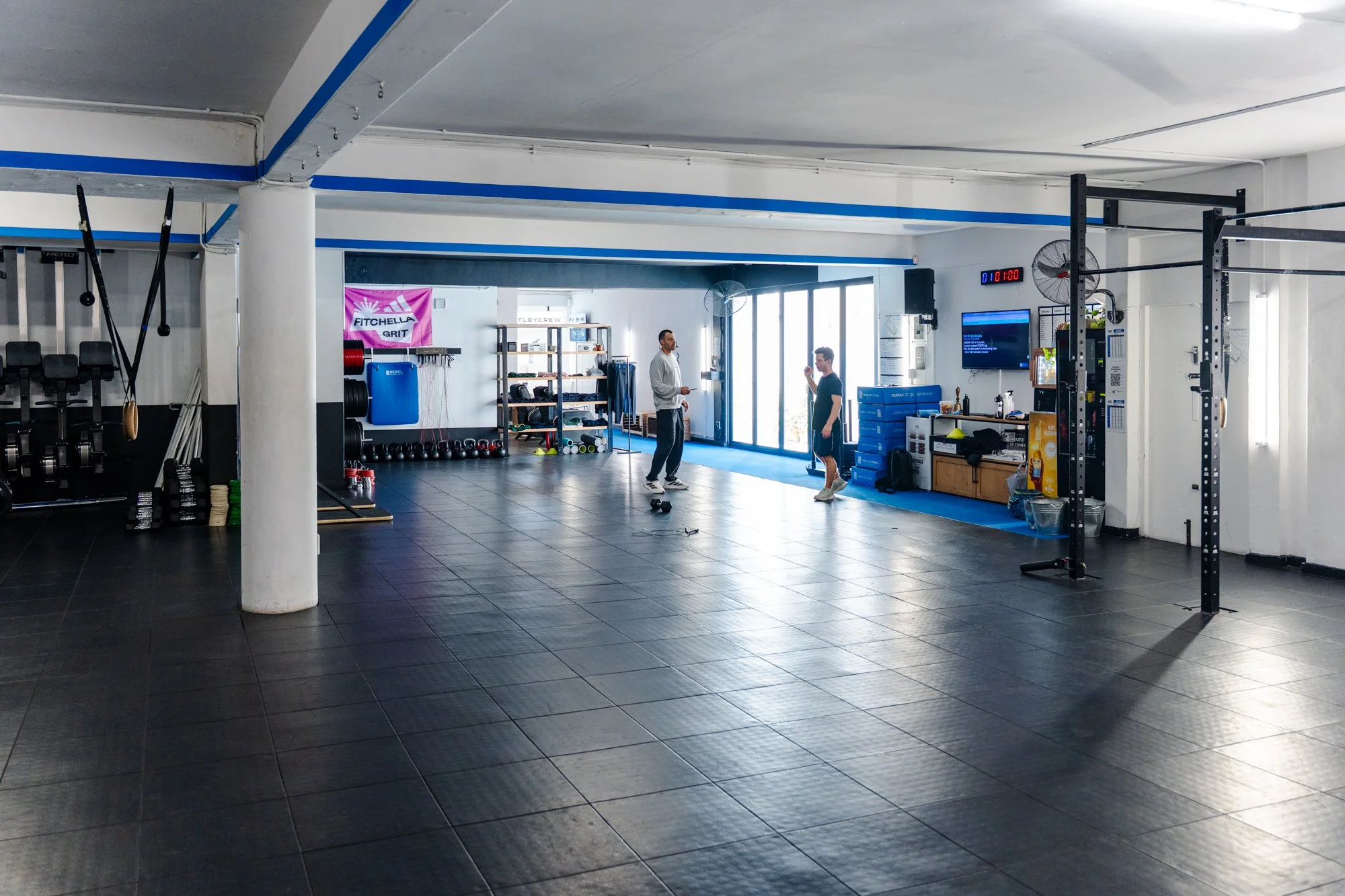Interior of a gym with black tile flooring, displaying two men having a conversation near a large window. The gym contains workout equipment, shelves with accessories, and a digital clock on the wall.