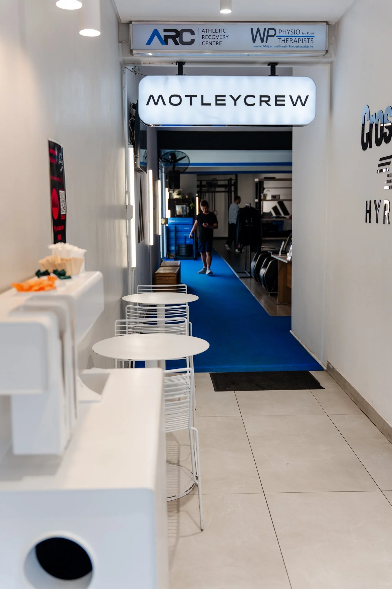 Indoor hallway leading to a fitness or physiotherapy center, with signs displaying ARC Athletic Recovery Centre and MOTLEYCREW, white tables and chairs in the foreground, and people walking inside.