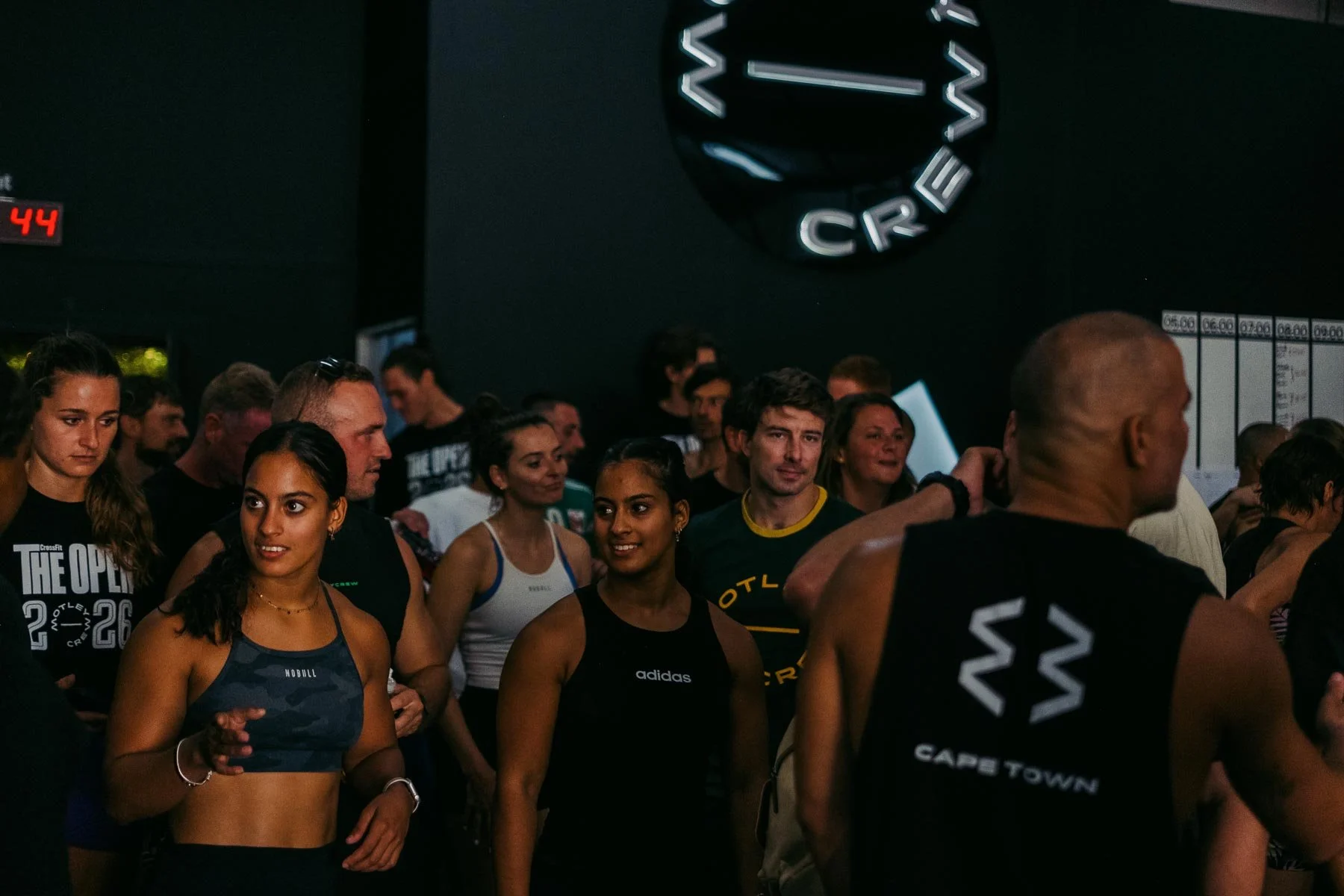 A group of people gathered at CrossFit event, some wearing athletic wear with the CrossFit logo or names, standing indoors with a dark wall, illuminated by a circular logo, and a scoreboard or schedule in the background.