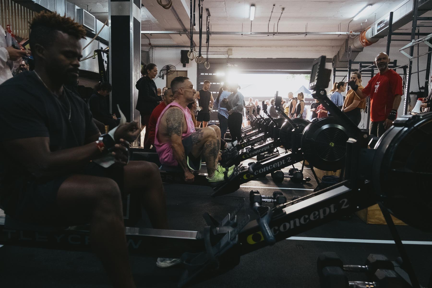 People working out on rowing machines inside a gym, with some people standing and observing, and others sitting or waiting. Bright sunlight is coming through the window.