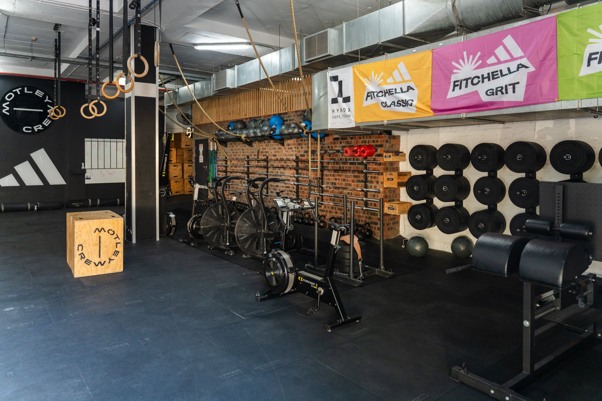 Interior of a gym with various workout equipment including rowing machines, medicine balls, barbells, and kettlebells. There are colorful banners for Atchelle, Fitchella Grit, and other brands hanging from the ceiling, with a brick wall and gym equipment on the back wall.