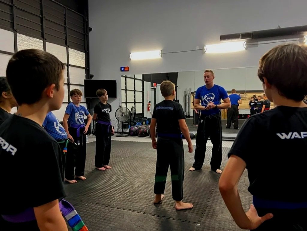 A medical or martial arts class where a young boy and an adult man, both dressed in black uniforms, are practicing martial arts with a focus on belt tying or a similar technique. Several people in the background, some wearing uniforms and some in casual clothing, watch or prepare for their turn.