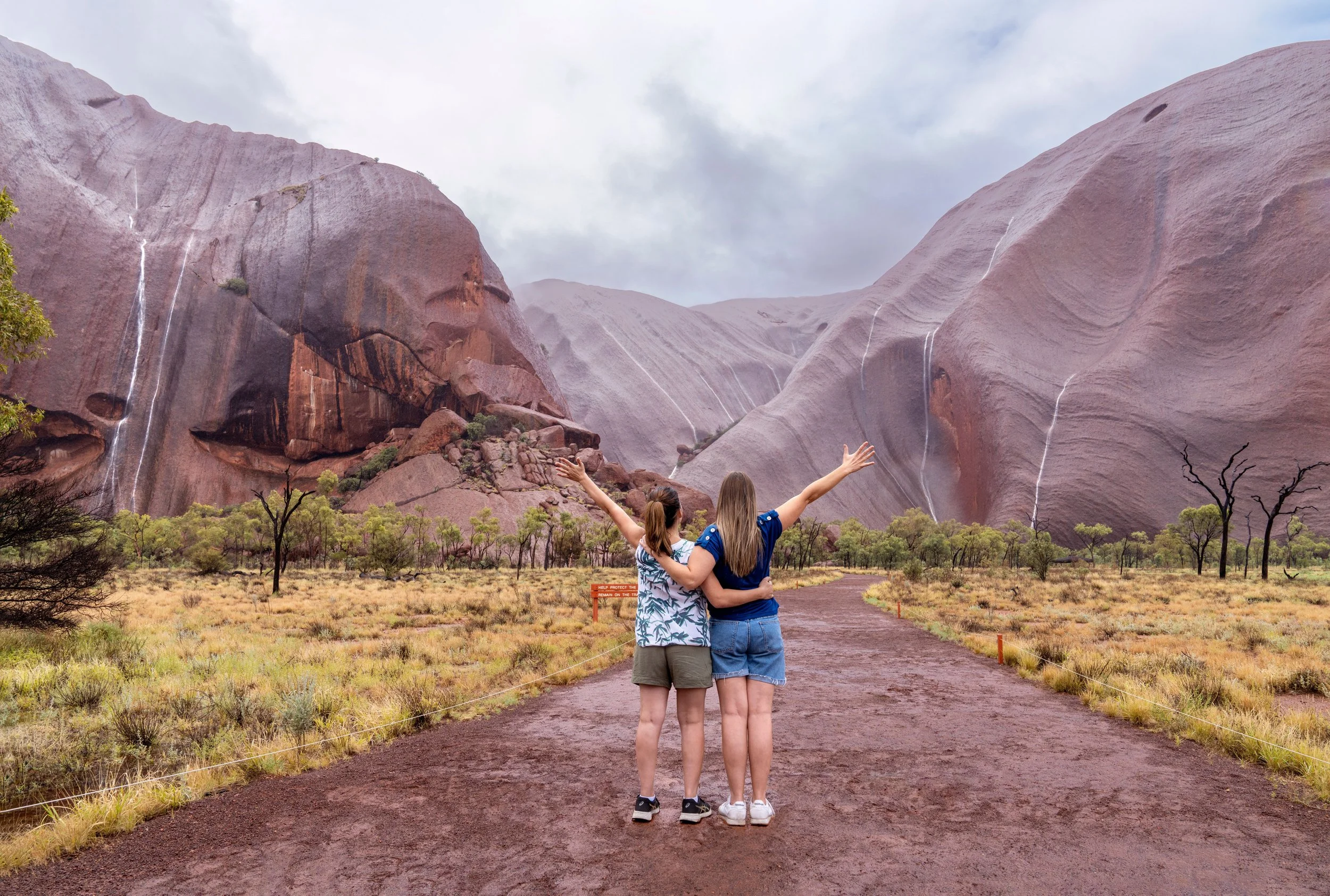 Two women standing on a dirt path with arms raised, facing large pinkish-purple rock formations of Uluru and waterfalls in a desert landscape with sparse trees and cloudy sky.