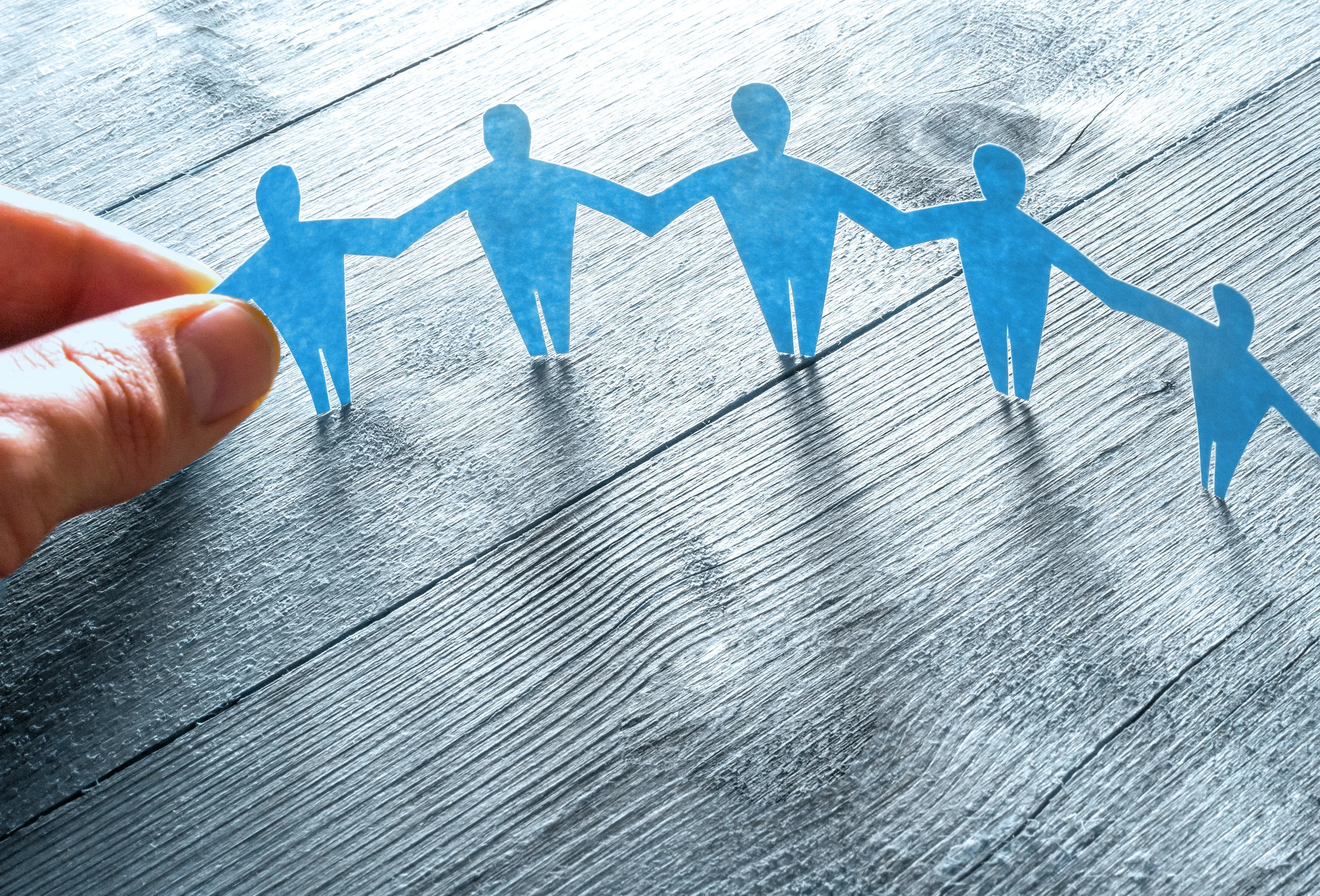 A hand holding a strip of blue paper cutouts of people holding hands, arranged in a circle on a gray wooden surface.