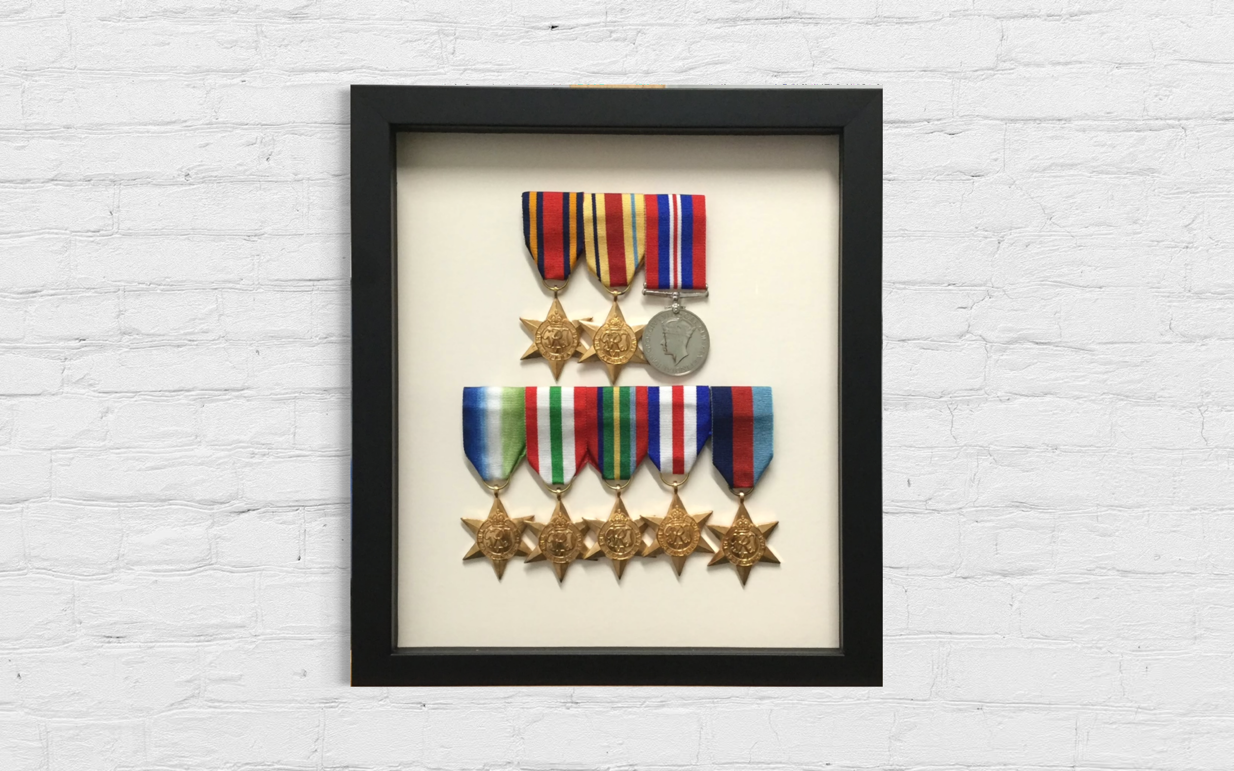 A framed display of military medals on a white background, featuring two gold medals with star-shaped designs and one silver medal with a profile of a soldier, all with colorful ribbons.