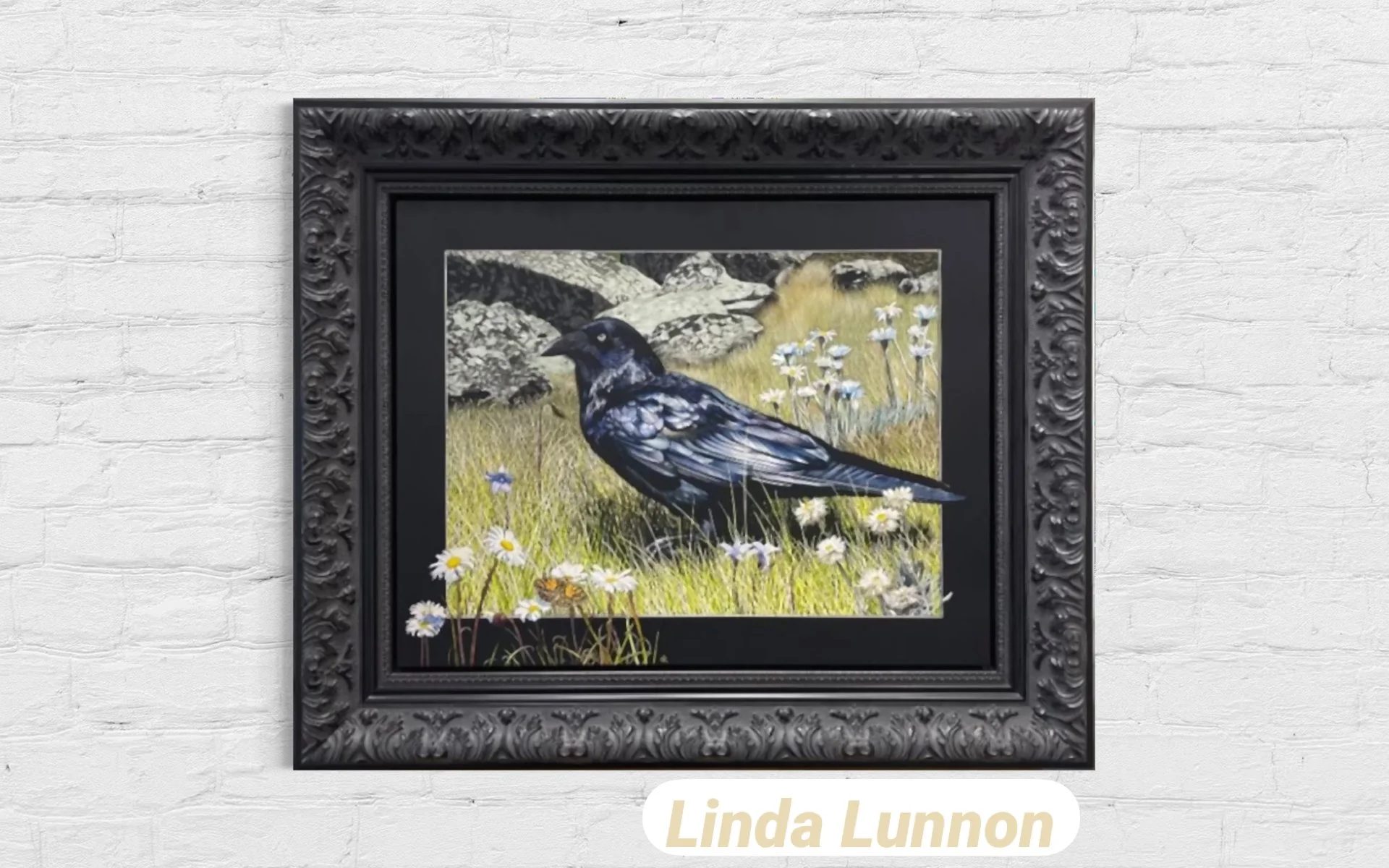A framed painting of a black bird standing in a grassy field with rocks and white daisies, set against a background of more rocks and grass.