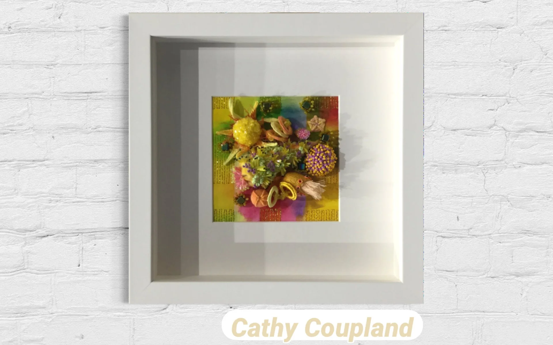 A white shadow box with a colorful floral arrangement inside, placed on a wooden surface against a brick wall background.