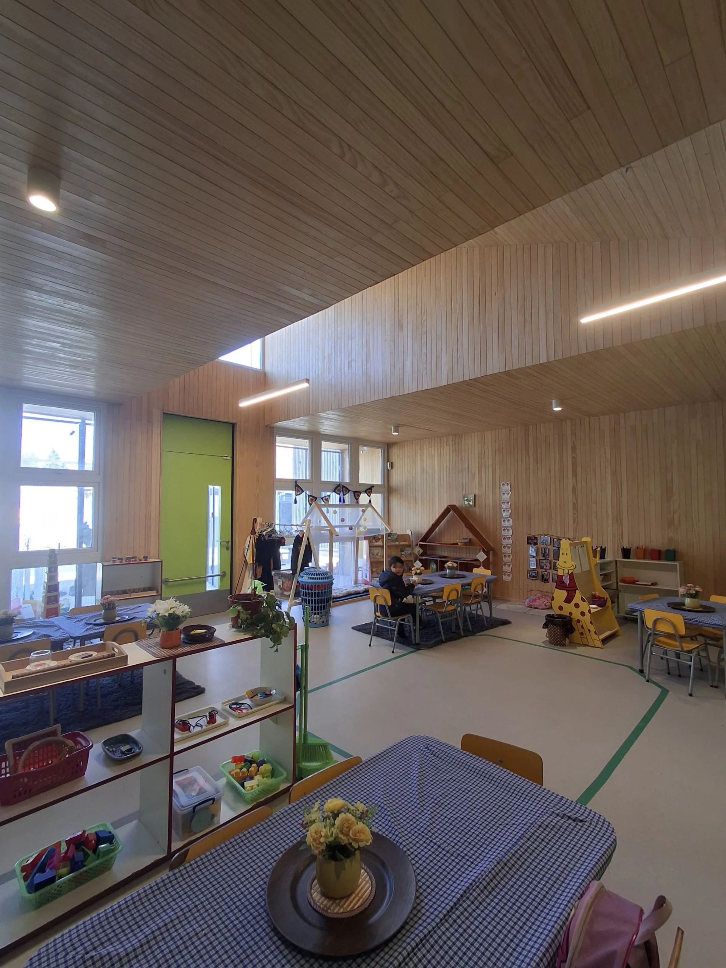 Interior de una sala de clases o área de actividades infantiles con paredes y techos de madera, varias mesas con manteles de cuadros, flores y juguetes, y ventanas que dejan entrar luz natural.