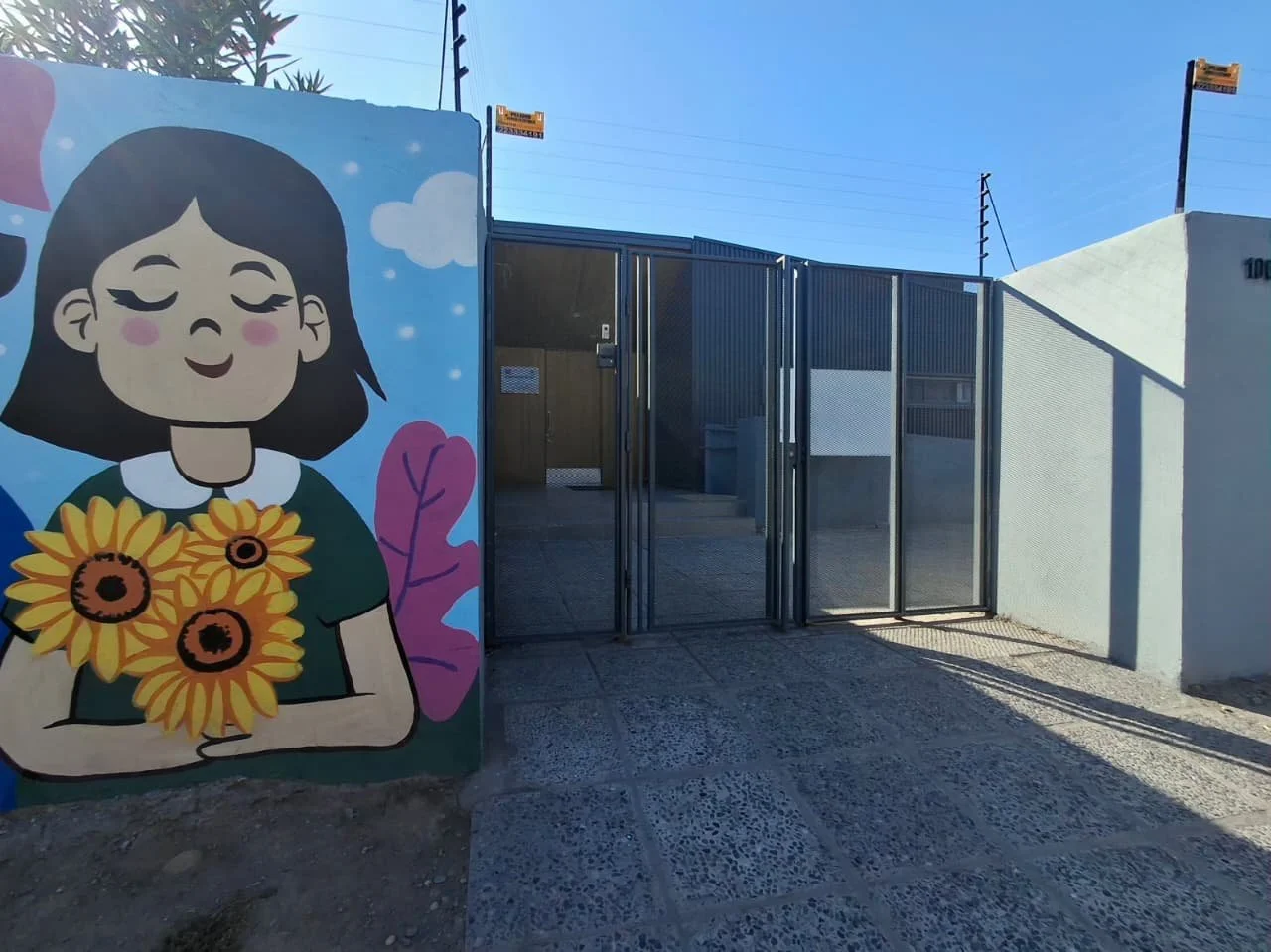 Mural colorido de una niña con cabello corto y ojos cerrados sosteniendo un ramo de girasoles, en un fondo azul con nubes y hojas rosas, junto a reja de entrada a un espacio cerrado