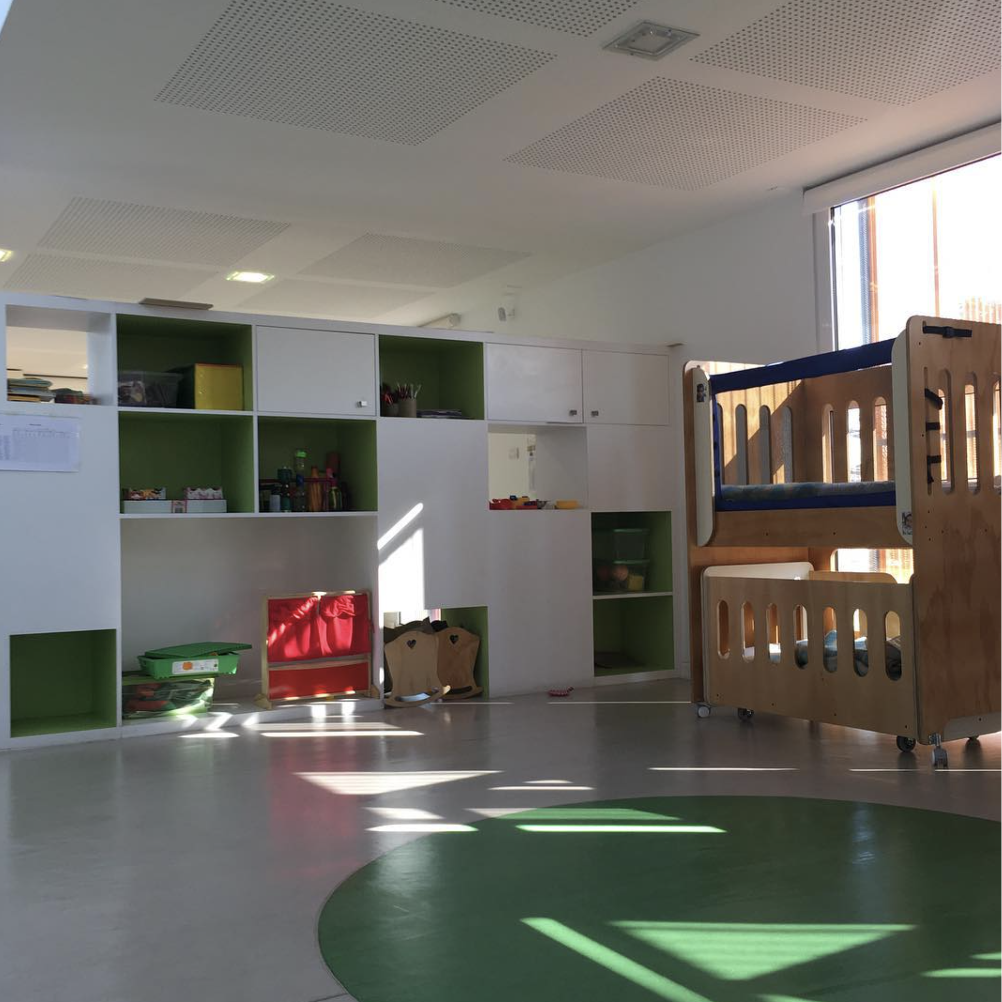 Sala de juego infantil con estanterías blancas y verdes, y un parque de juegos de madera con ruedas.