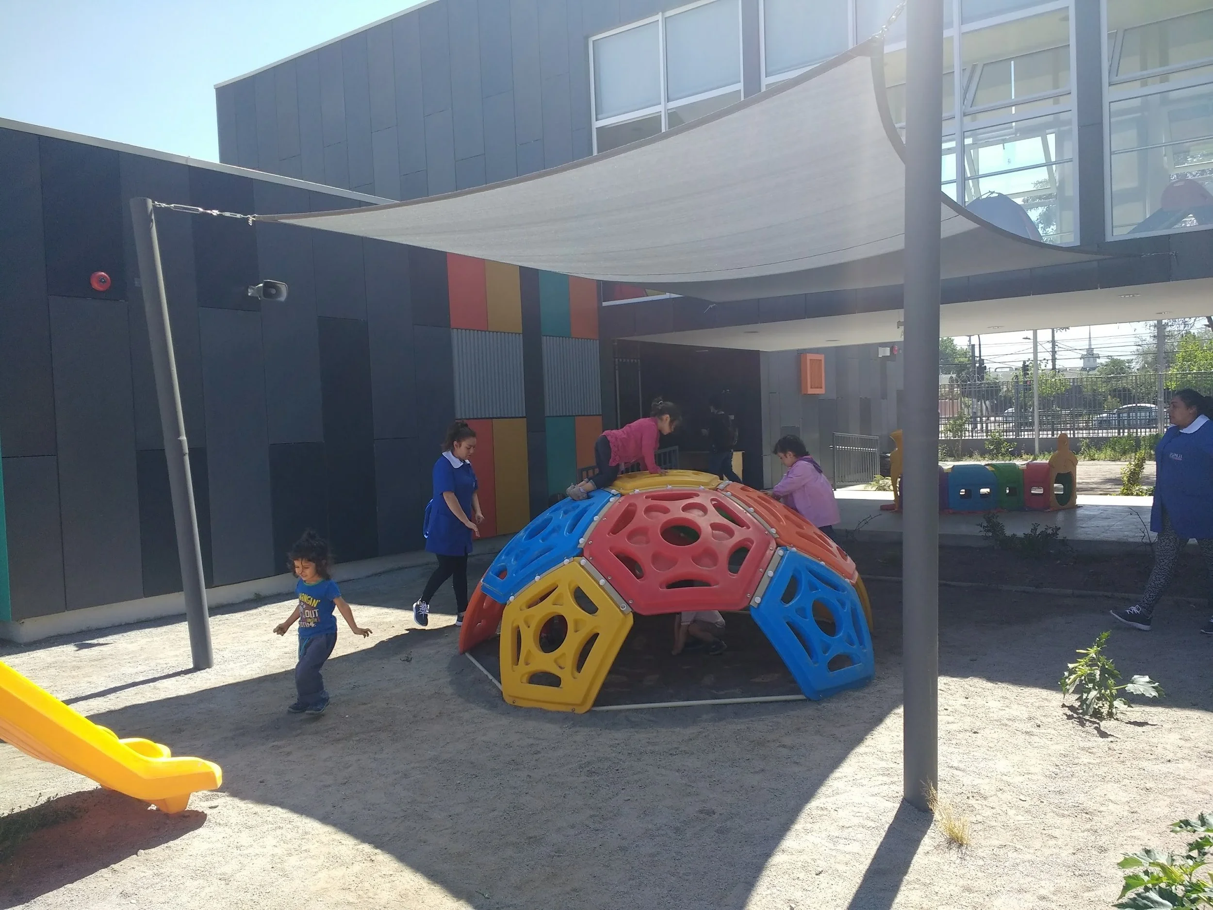 Área de juegos infantiles con estructuras de plástico en colores amarillo, azul, naranja y verde, y personas adultas y niños jugando bajo sombra de una lona blanca en un espacio exterior, con edificio y alambrado de fondo.