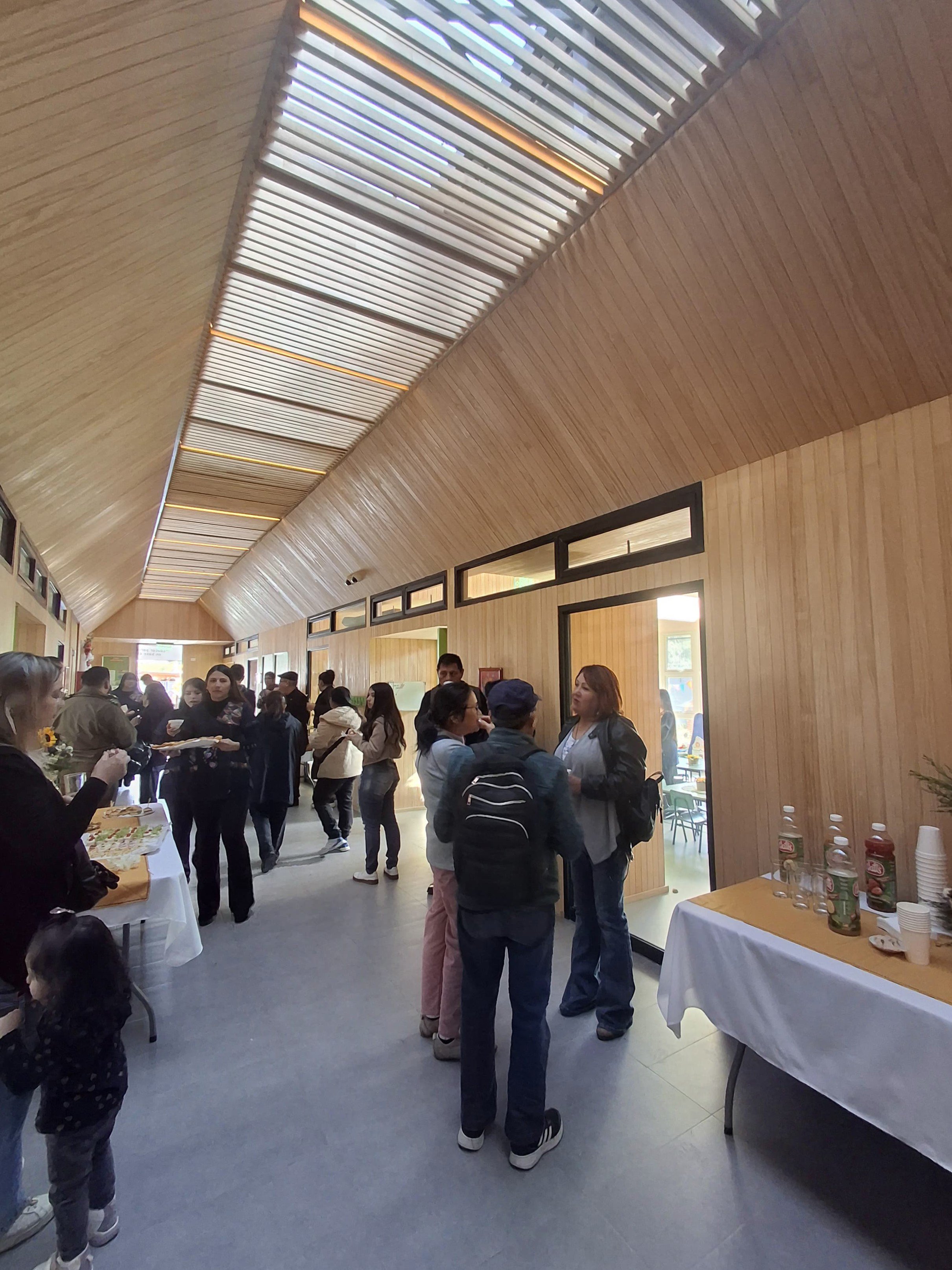 Vista de interior con muchas personas en una reunión o evento, paredes de madera clara y techo con estructuras de color beige y líneas de luz, mesas con comida y bebidas.