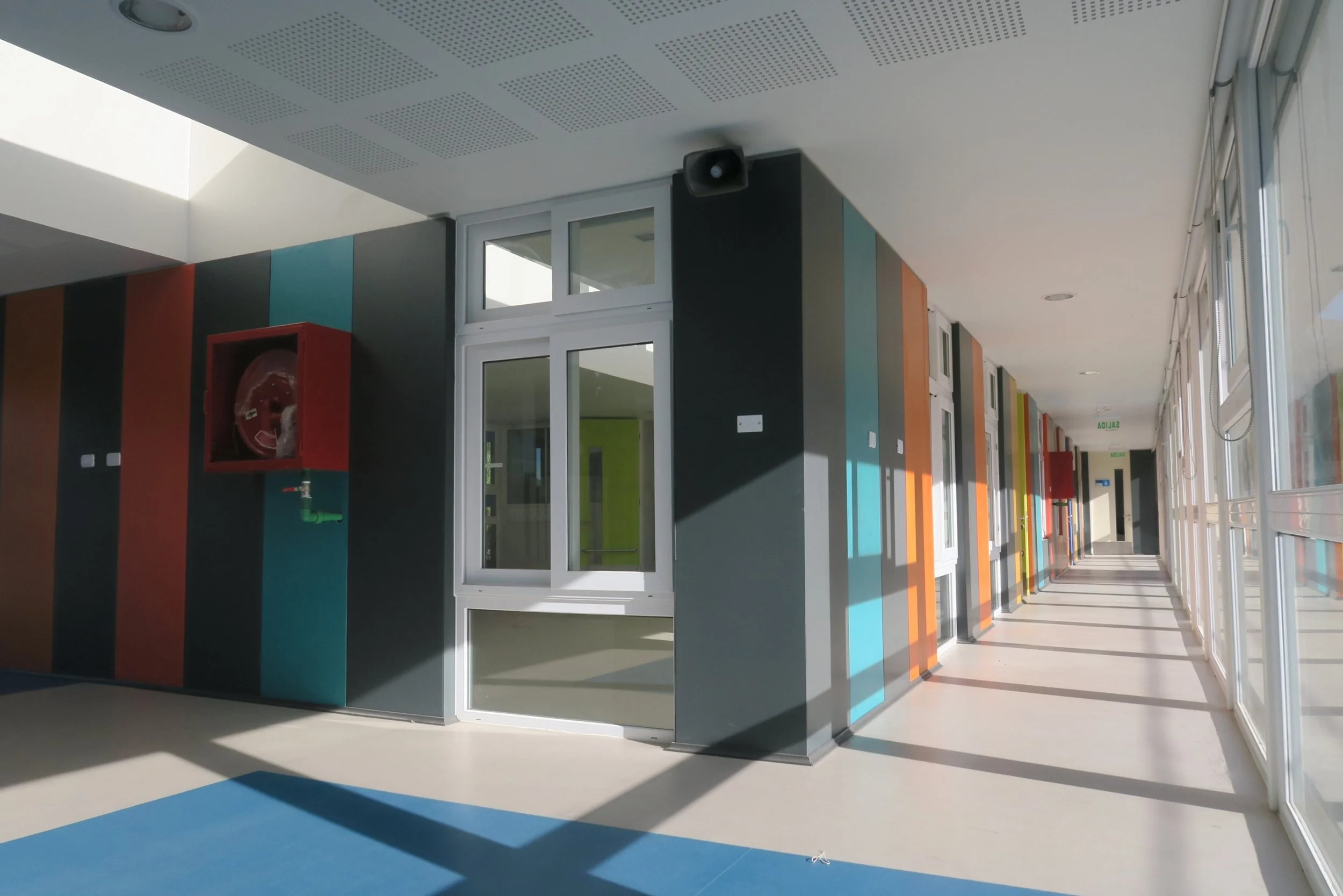 Pasillo largo en un edificio moderno con paredes decoradas con franjas de colores y ventanas grandes por donde entra luz natural, incluyendo conductos de aire en el techo y un extintor en una pared roja.