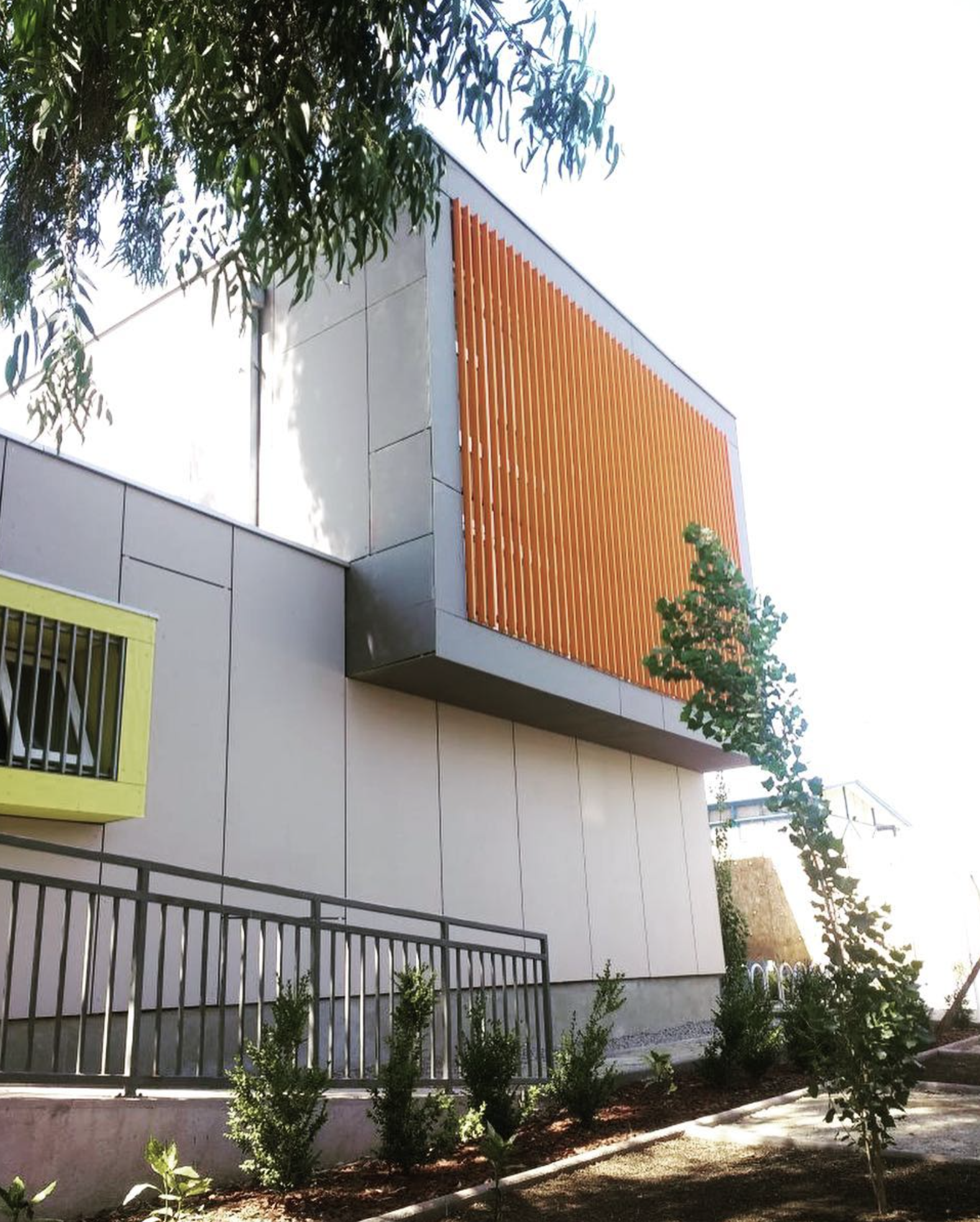 Edificio moderno con paredes de color gris y un gran ventanal con rejas amarillas. La fachada principal tiene un diseño con listones de madera y jardineras con arbustos y árboles pequeños en el jardín frontal.