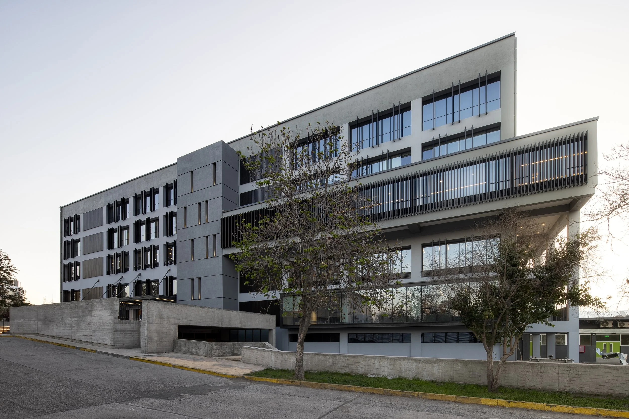 Edificio moderno de varios pisos con ventanas grandes y persianas negras, árboles sin hojas en primer plano, y una acera en un entorno urbano.