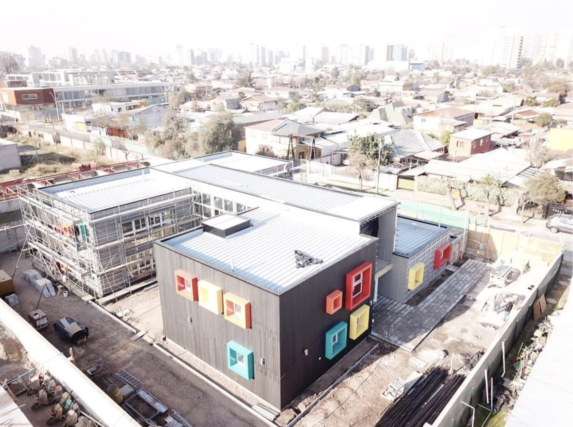 Construcción de un edificio moderno con paredes de color gris y ventanas decoradas con marcos en diferentes colores, en un barrio urbano. Tiene una terraza y estructuras de andamios en un lado, y se observa el vecindario en el fondo.