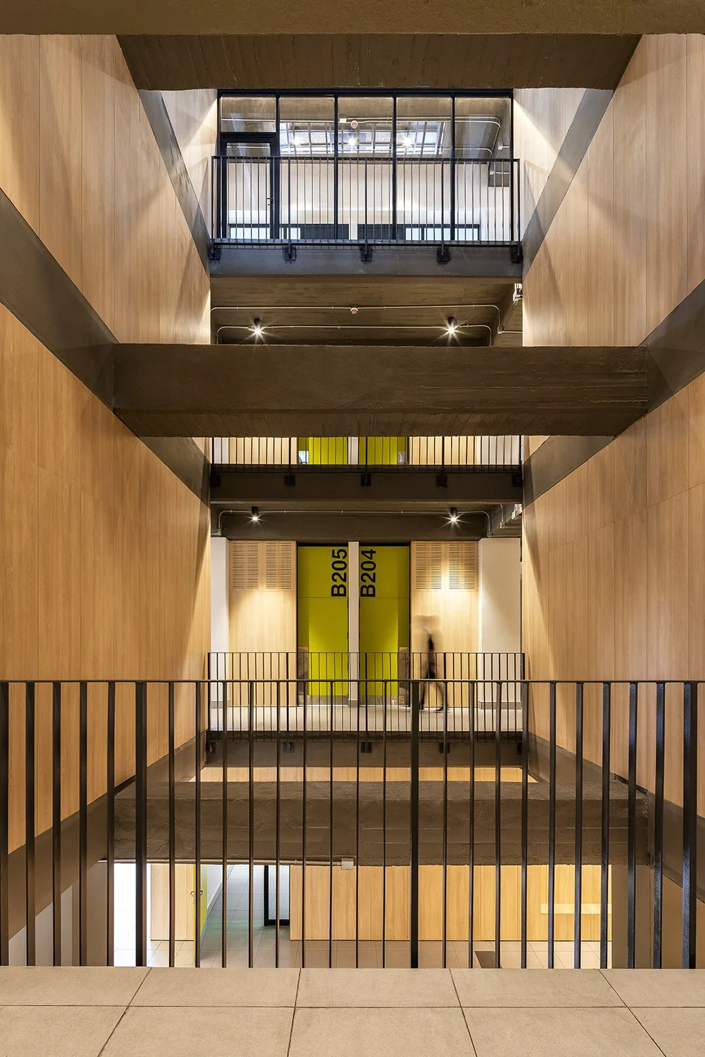 Vista interior de un edificio con varias plantas conectadas por pasarelas de concreto y barandales de metal, con paredes de madera y puertas de colores con etiquetas B205 y B204.