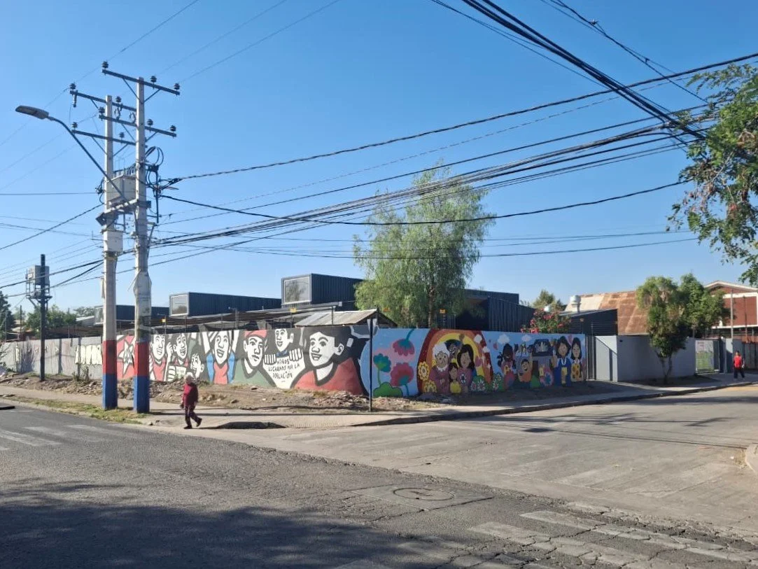 Mural colorido de rostros sonrientes, flores y personajes infantiles en una cerca en una calle urbana con cables y árboles