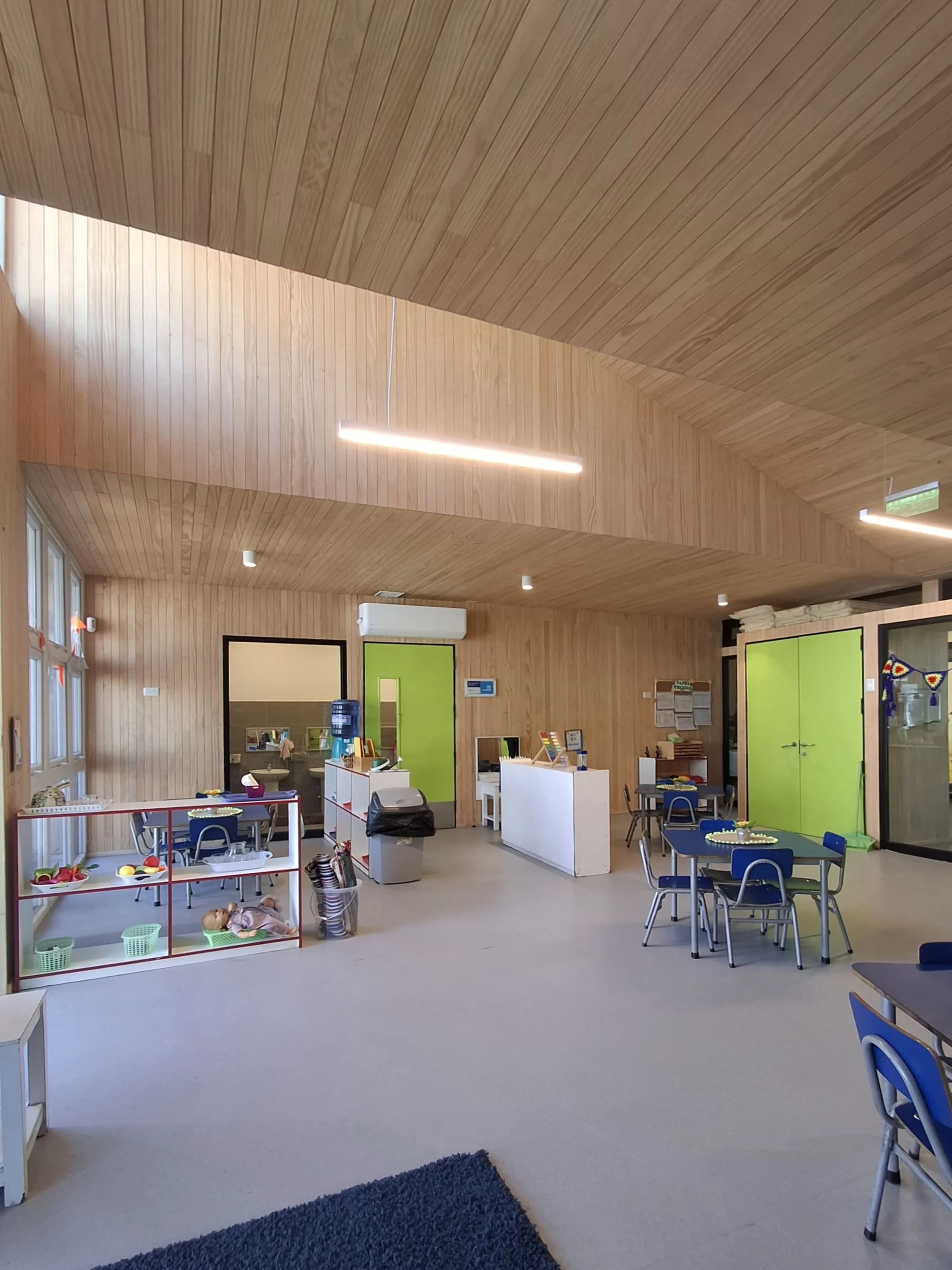 Interior de un aula de jardín de niños con paredes y techo de madera, varias mesas y sillas, zona de juegos con muñecos y juguetes, equipo de aire acondicionado y ventanas grandes que dejan entrar luz natural.