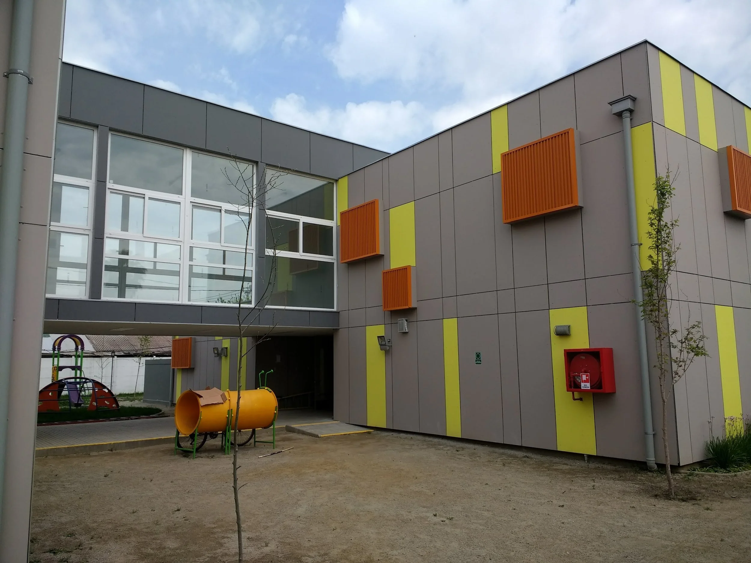 Edificio escolar modernizado con ventanas grandes, paredes decoradas con bloques de color gris y amarillo, y varias estructuras en el patio infantil, incluyendo un túnel de juego naranja y un columpio en forma de media luna de colores.»