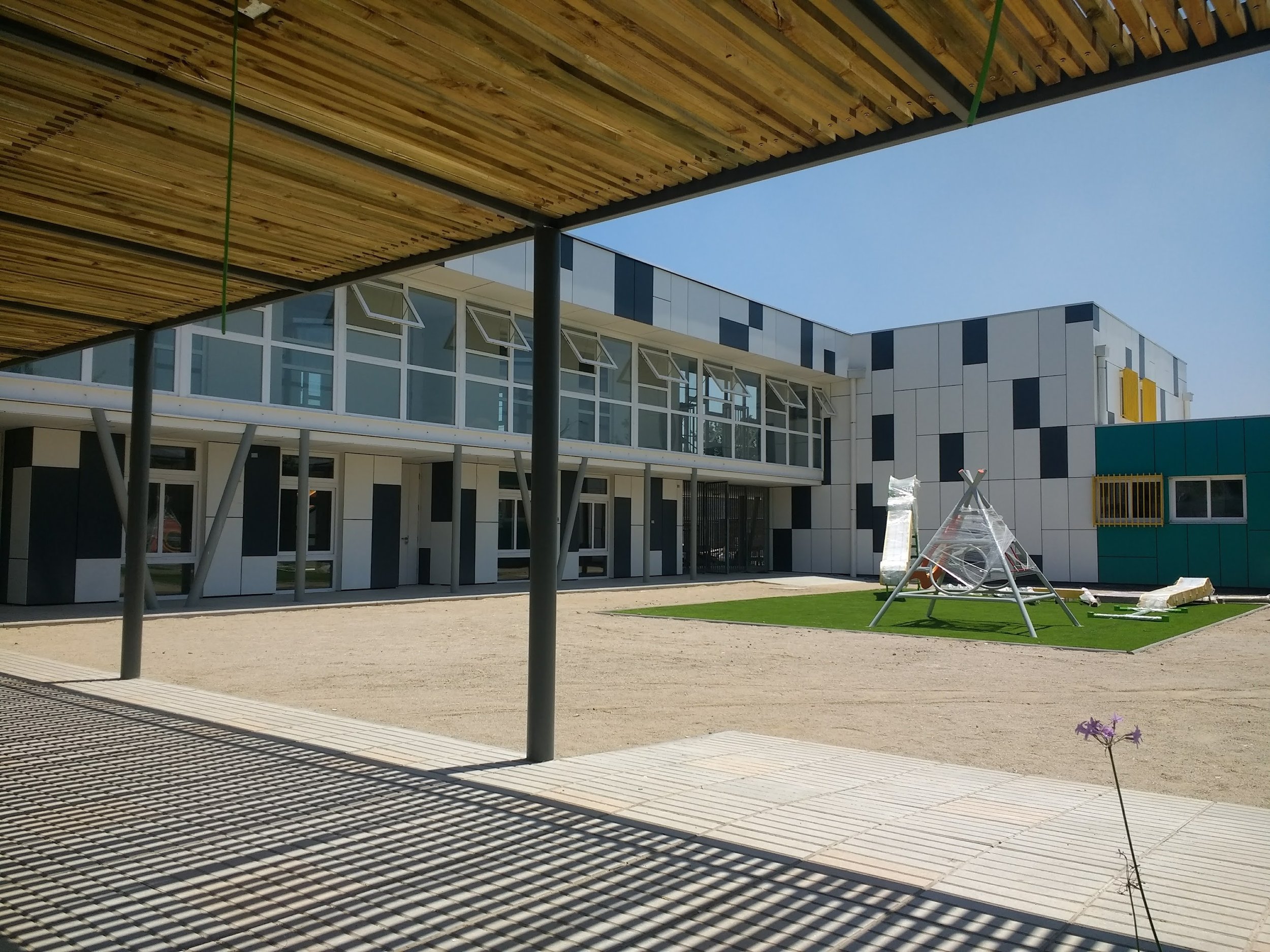 Vista de un edificio moderno con ventanas grandes y un diseño en cuadrícula de colores blancos, negros y amarillos. En el exterior hay una estructura de sombra hecha de madera en forma de techo y un columpio en forma de cabaña. También hay un área de
