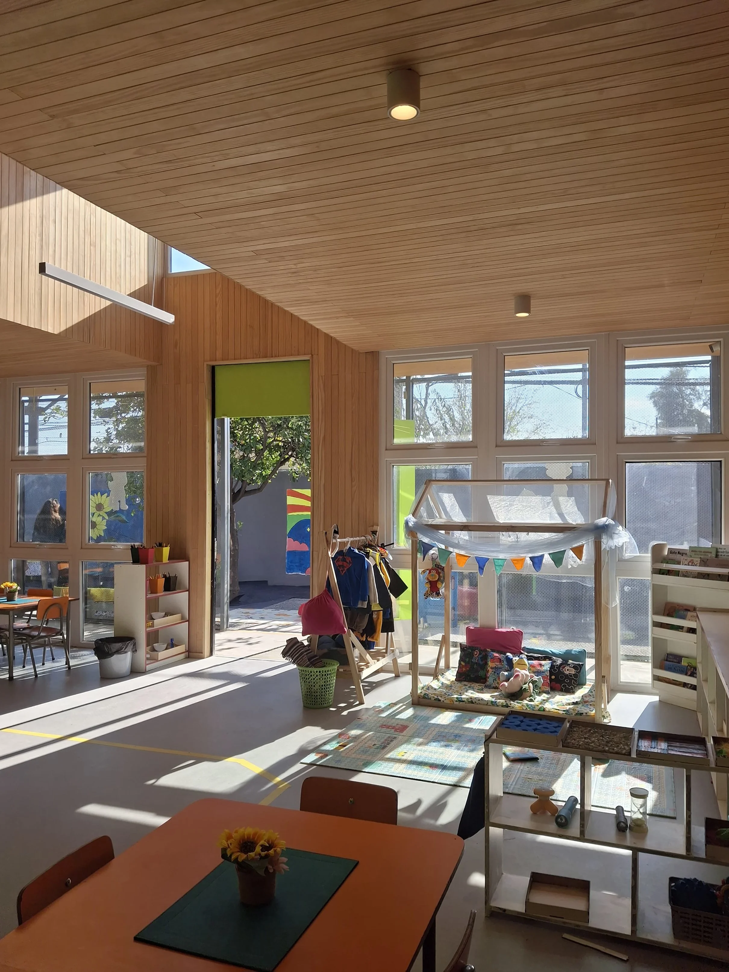 Interior de una aula escolar con ventanas grandes, techo de madera, y área de juegos y libros. Hay mesas y sillas, decoración colorida, y objetos de aula.