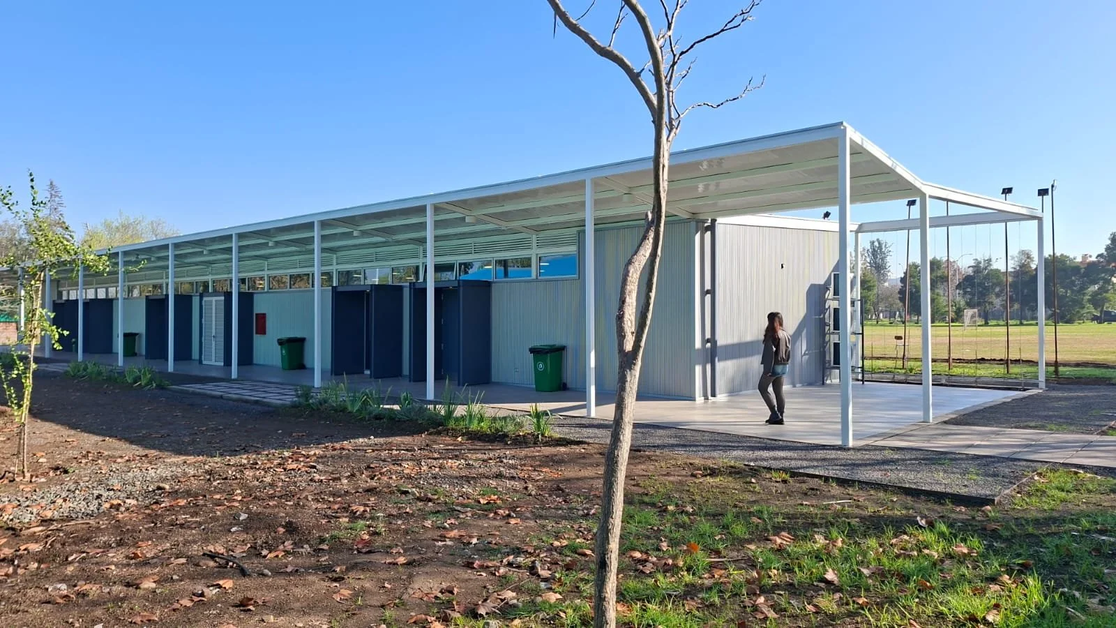 Edificio de estructura moderna con paredes cubiertas de plantas verdes, en un parque con césped, arbustos y flores moradas, con varias personas paseando y jugando, y ave volando en el cielo azul con nubes.