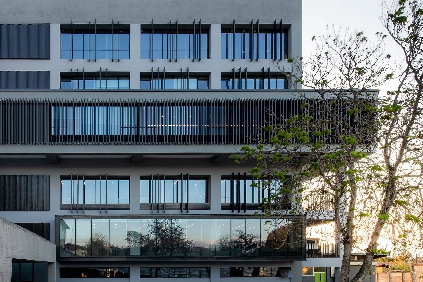 Edificio moderno de varios pisos con balcones y ventanas de vidrio, con árboles alrededor y atardecer al fondo.