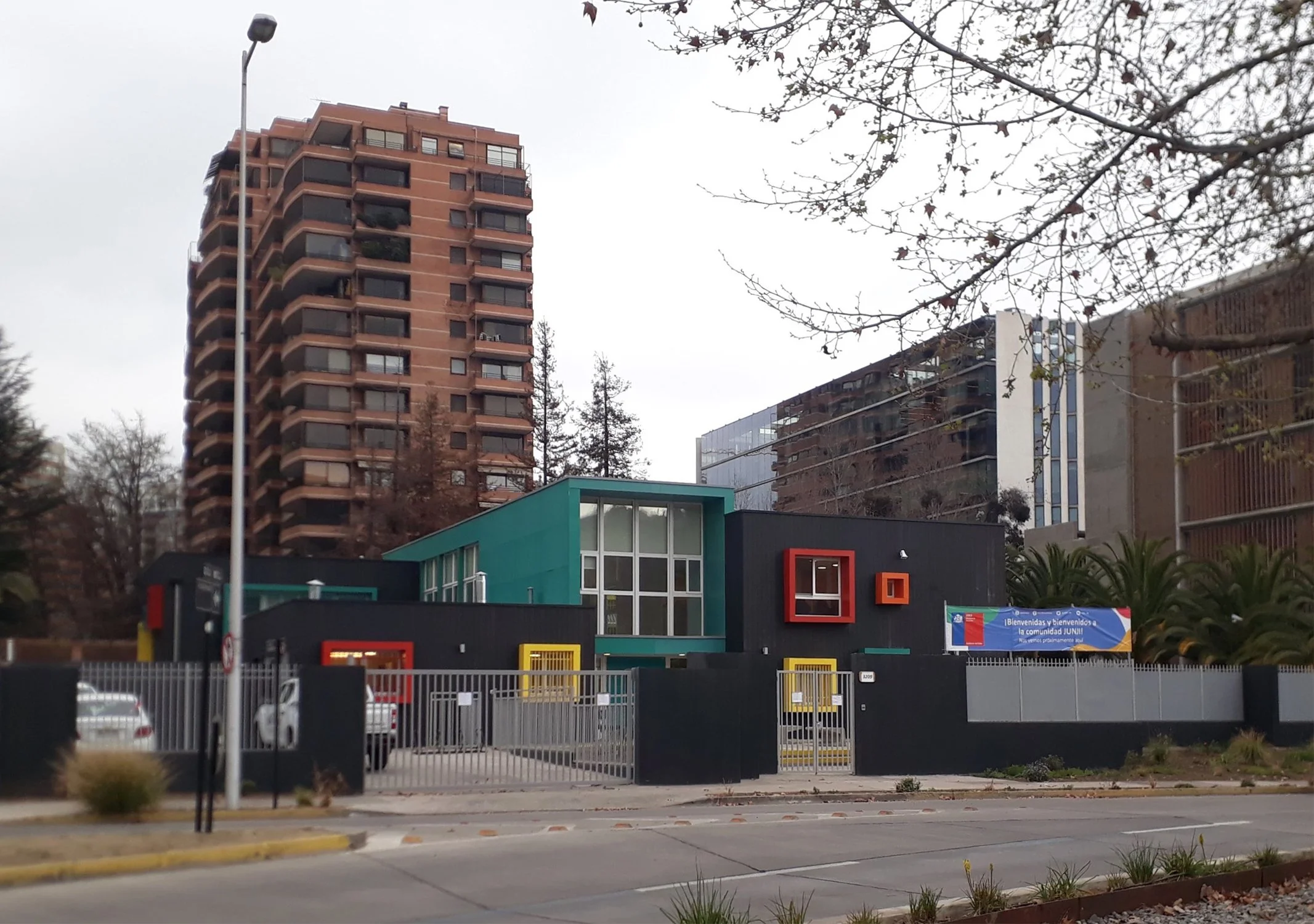 Edificio de,color negro y verde con ventanas grandes y decoraciones en colores rojo y amarillo, con valla de metal en la parte frontal y árboles en el fondo.