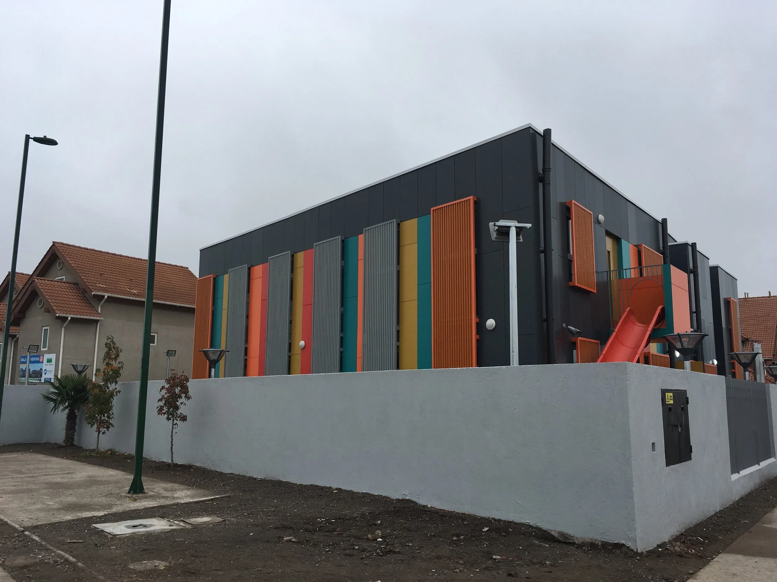 Edificio moderno con paredes decoradas con paneles de colores y un tobogán naranja, en un área con lámparas de luz y algunos árboles pequeños, bajo cielo nublado.